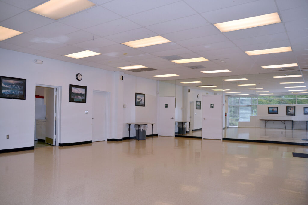 Empty room with polished floor, fluorescent ceiling lights, wall mirrors, framed pictures, a clock, and doors leading to adjoining rooms. Tables are visible through the far windows.