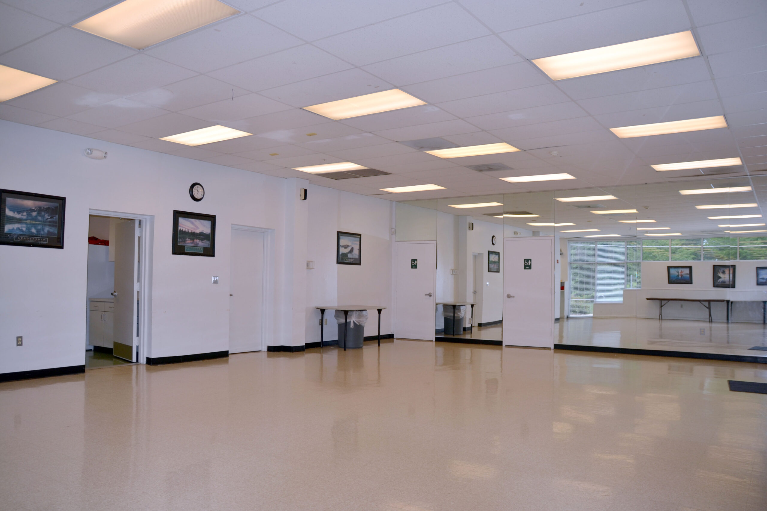 Empty room with polished floor, fluorescent ceiling lights, wall mirrors, framed pictures, a clock, and doors leading to adjoining rooms. Tables are visible through the far windows.
