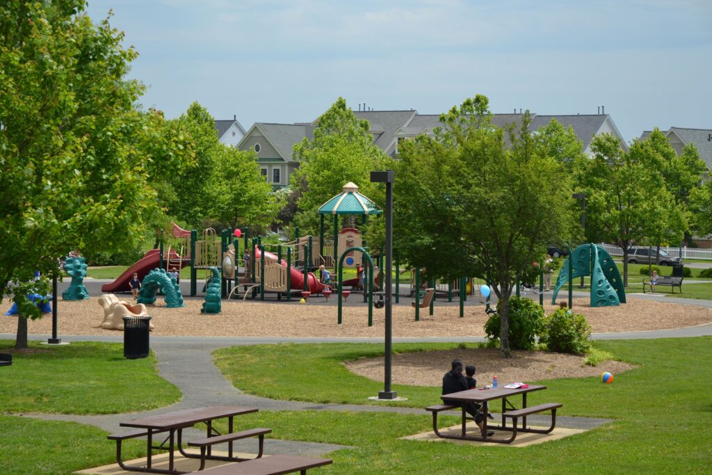 A playground with slides and climbing structures is surrounded by green trees. Picnic tables and a person sitting on a bench are in the foreground; houses are in the background.