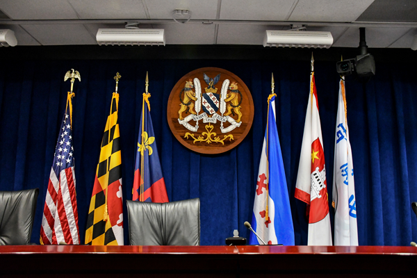 A government press conference room with five flags, leather chairs, and a crest mounted on a dark blue curtain backdrop.