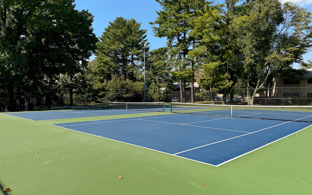 Two empty blue and green outdoor tennis courts surrounded by trees under a clear sky.