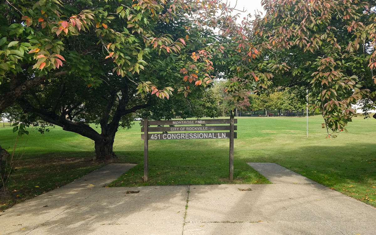 A wooden park sign for Montrose Park, 451 Congressional Ln, City of Rockville, stands between two trees by a grassy field.