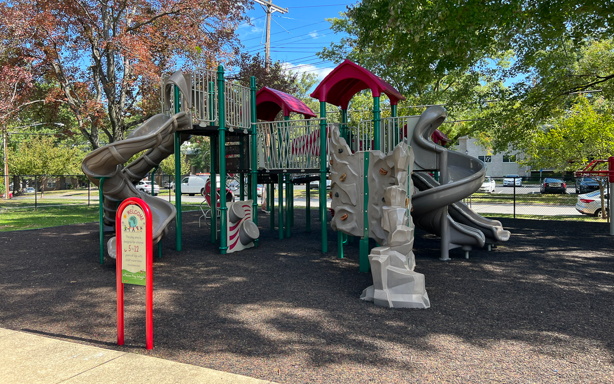 A playground with slides, climbing walls, and overhead bridges stands on a rubber surface surrounded by trees and parked cars on a sunny day.
