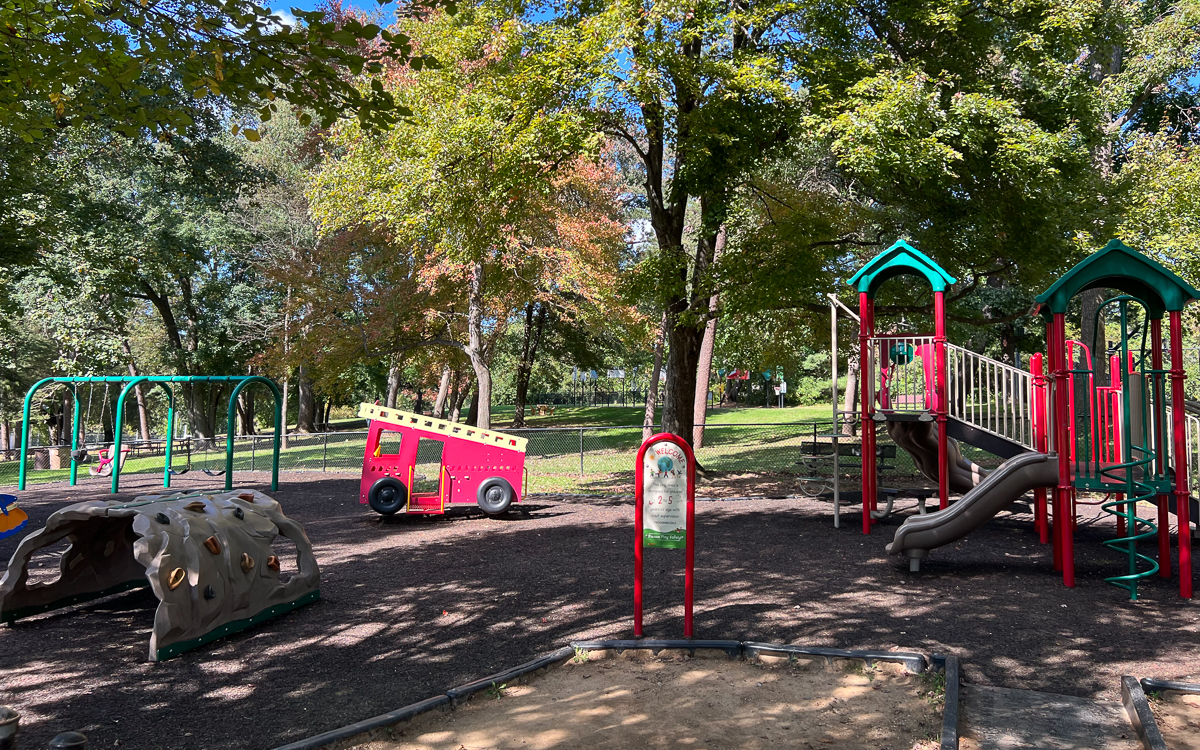 Children’s playground with slides, climbing structures, swings, and a red play vehicle surrounded by trees and greenery on a sunny day.