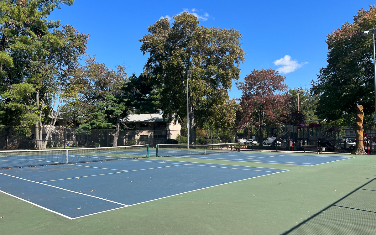 Two empty outdoor tennis courts surrounded by trees and a fence on a sunny day with a clear blue sky.