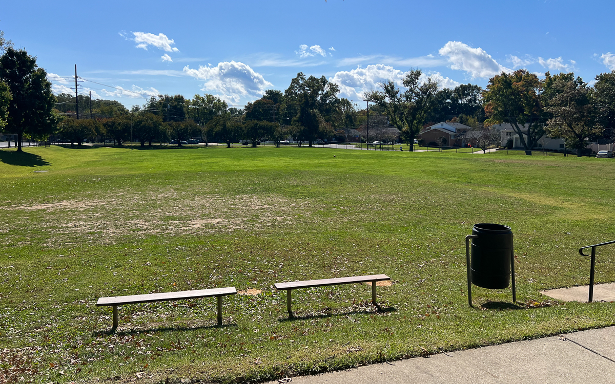 A grassy park field with two benches and a trash can in the foreground, trees and houses in the background under a blue sky with clouds.