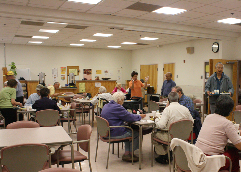 People of various ages sit and eat or interact in a cafeteria-style room with tables, chairs, and food service counters in the background.