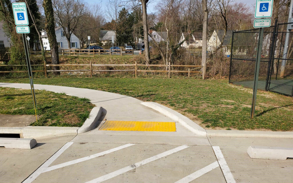 Concrete sidewalk with a yellow textured ramp connects two accessible parking spaces to a grassy area, near a wooden fence and tennis court.