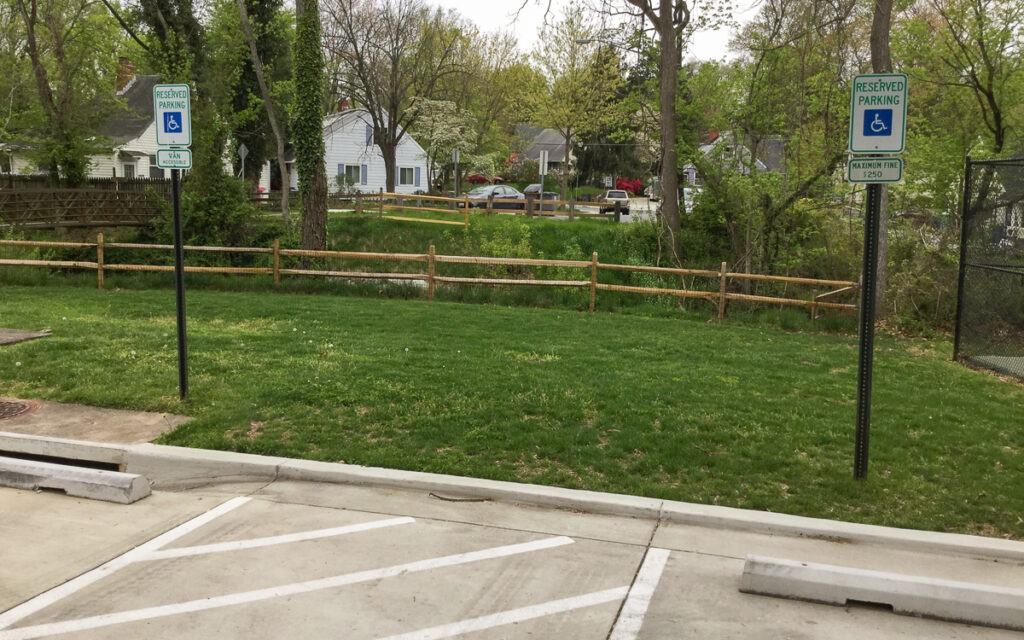 Two empty accessible parking spaces with reserved signs next to a grassy area, wooden fence, and trees in a residential neighborhood.