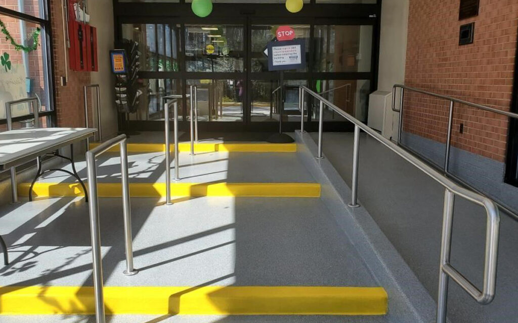 Entrance with automatic glass doors, three accessibility ramps with yellow curbs, handrails, and a stop sign with a notice posted on the door.