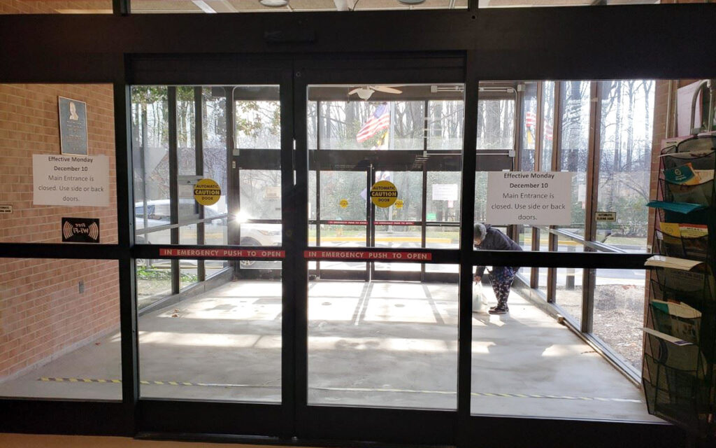 Glass double doors with caution tape and signs stating the main entrance is closed, advising use of side or back doors; a person is seen outside near the entrance.
