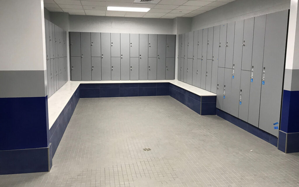 Empty locker room with gray lockers lining the walls, a tiled floor, and a white bench running along two sides of the room.
