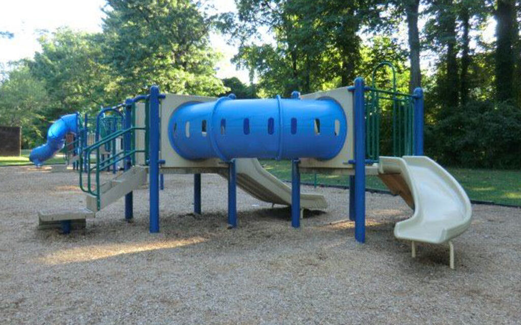 Playground structure with blue and beige slides, climbing steps, and a tunnel tube, set on a gravel surface and surrounded by trees.