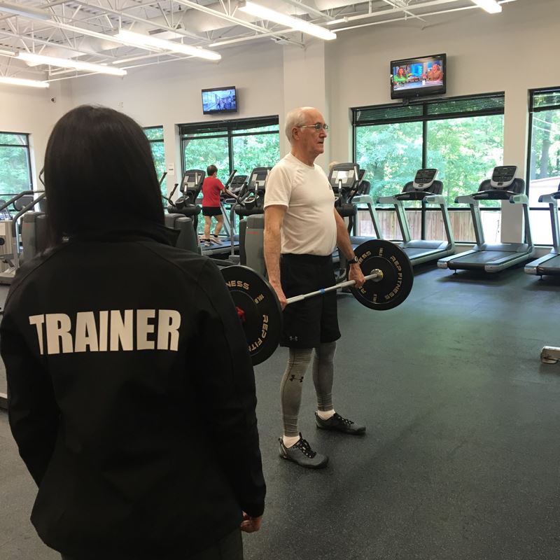 An older man lifts a barbell in a gym while a trainer observes. Treadmills and other people exercising are visible in the background.