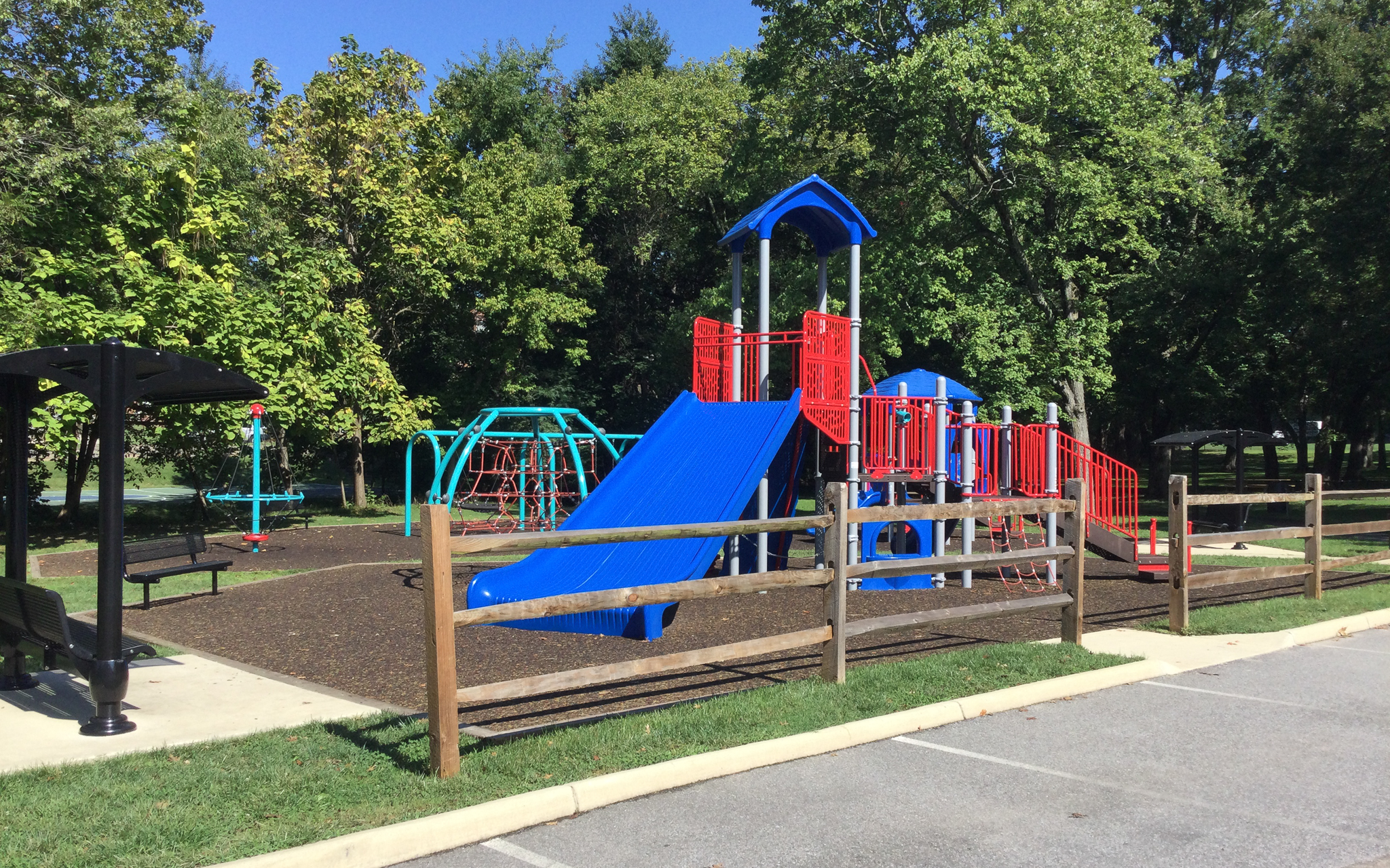 A playground with blue and red climbing structures and slides, a swing set, a bench under a canopy, and a wooden fence next to a parking area. Trees are in the background.