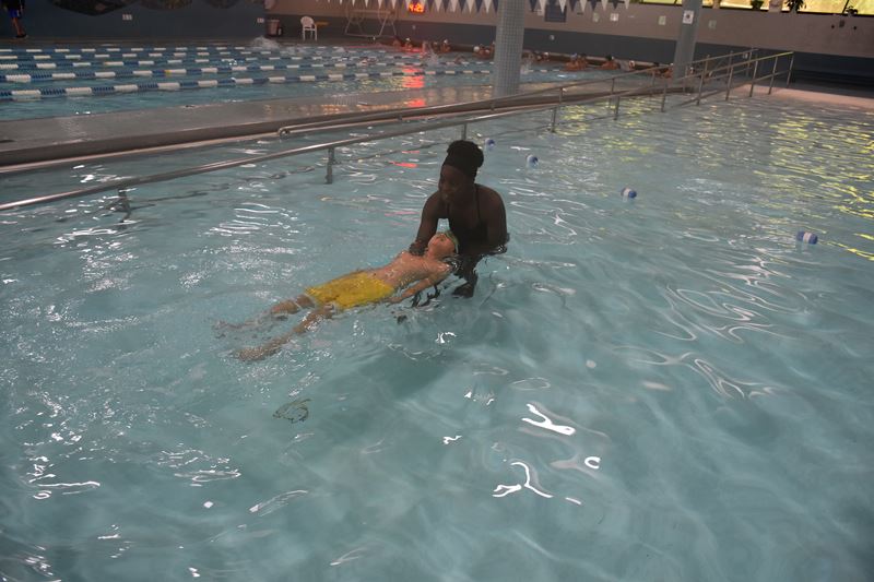 An adult helps a child in yellow shorts float on their back in a swimming pool during a swim lesson.