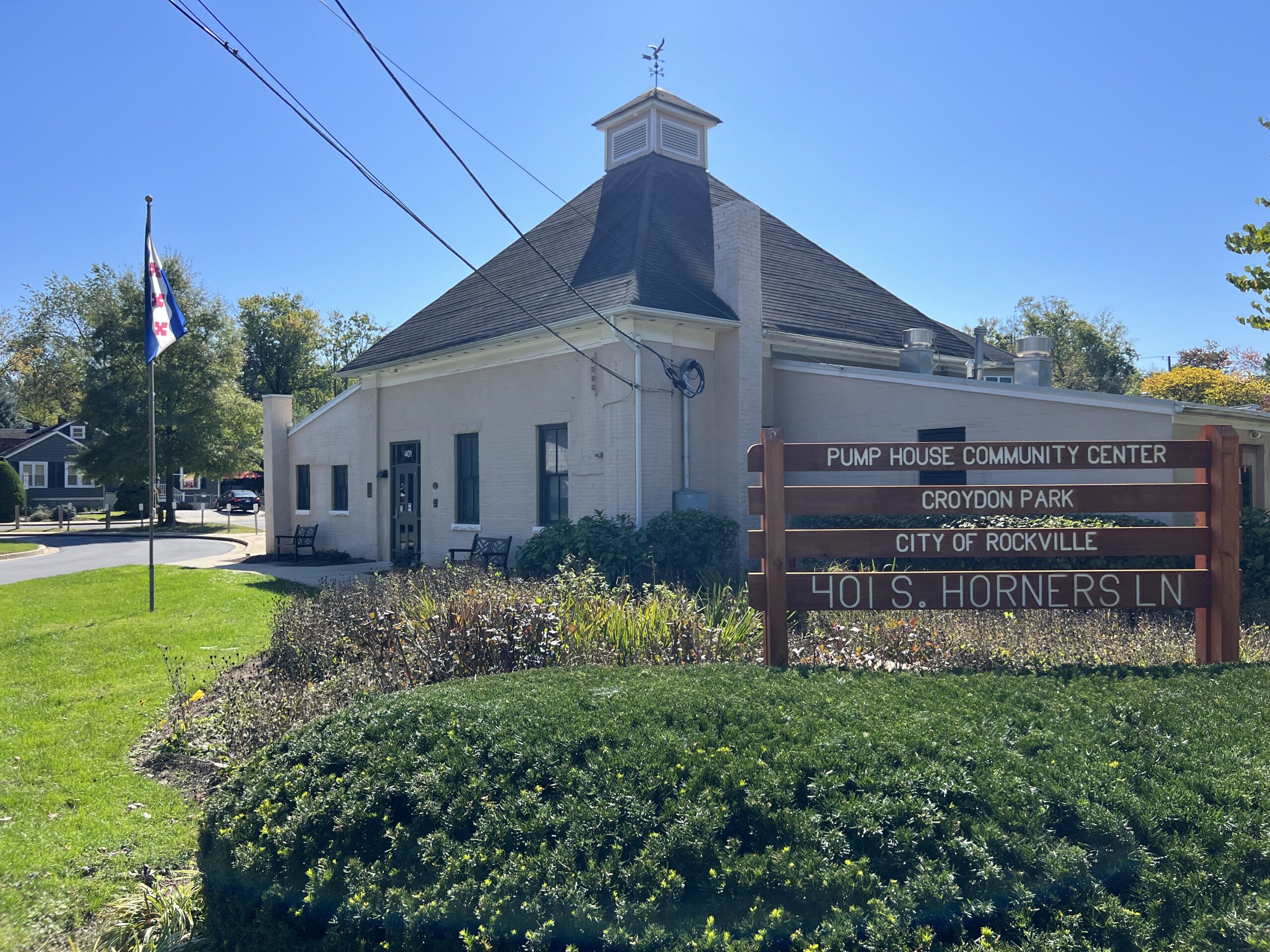 A beige building with a sign reading "Pump House Community Center, Croydon Park, City of Rockville, 401 S. Horners Ln" stands next to a flagpole and greenery on a sunny day.