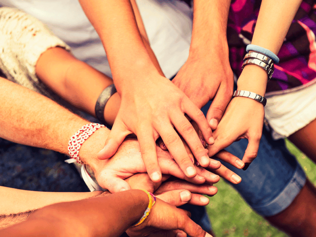 Several people stack their hands together in a gesture of unity and teamwork, with various bracelets and watches visible on their wrists.