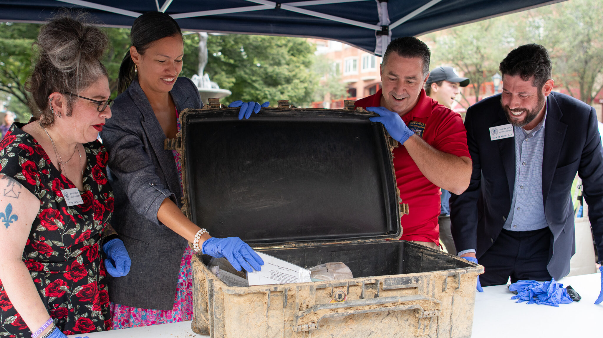 Four people wearing gloves examine documents inside a weathered, open suitcase on a table under a canopy during an outdoor event.