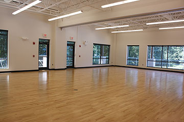 Spacious, empty room with light wood flooring, large windows, white walls, and ceiling lights, overlooking trees outside.