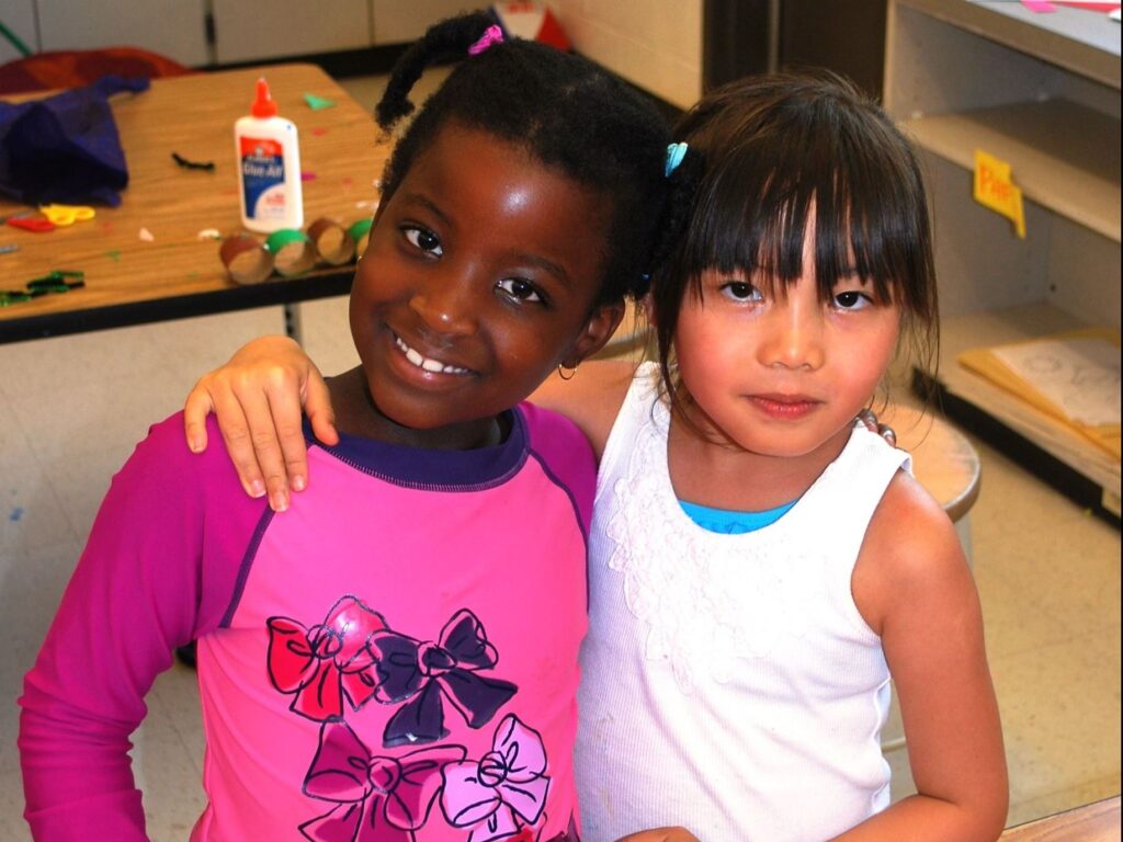 Two young girls stand side by side in a classroom, one with her arm around the other. Art supplies and papers are visible on a table and shelves in the background.