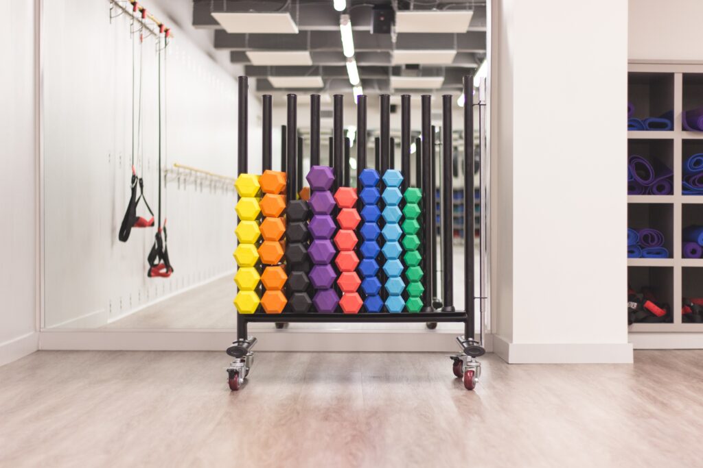 A rack of colorful hexagonal dumbbells is organized by weight in a gym, with exercise equipment and yoga mats in the background.