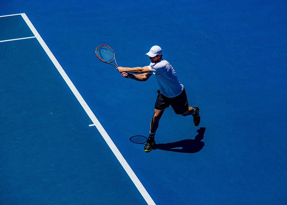 A tennis player wearing a white shirt and cap hits a backhand shot on a blue hard court.
