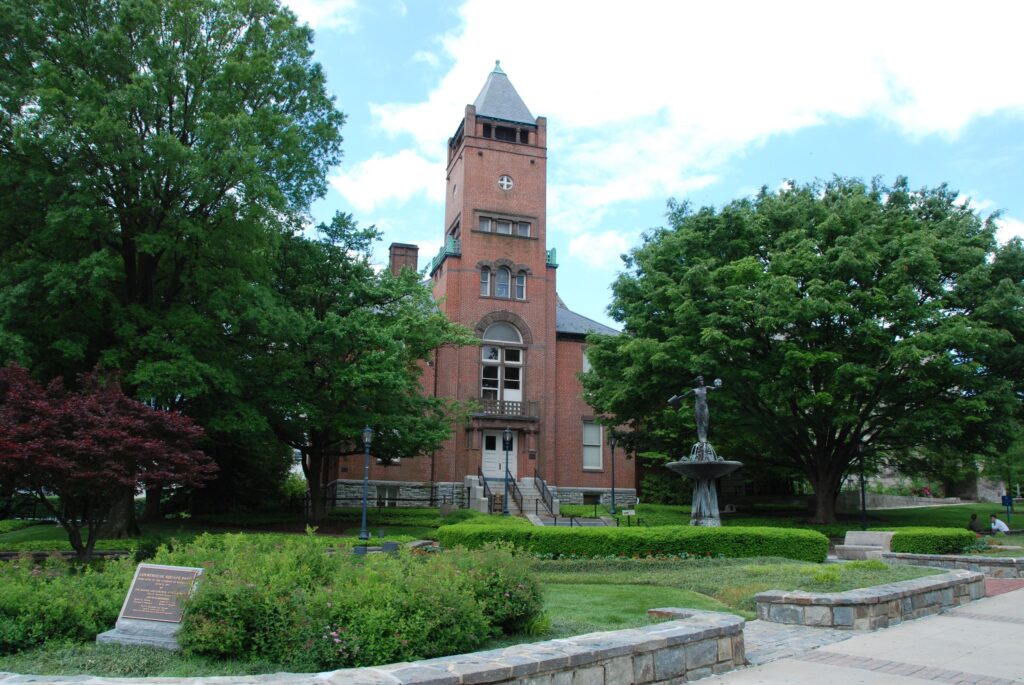 A historic red brick building with a clock tower stands behind a landscaped garden, large trees, and a stone border under a partly cloudy sky.