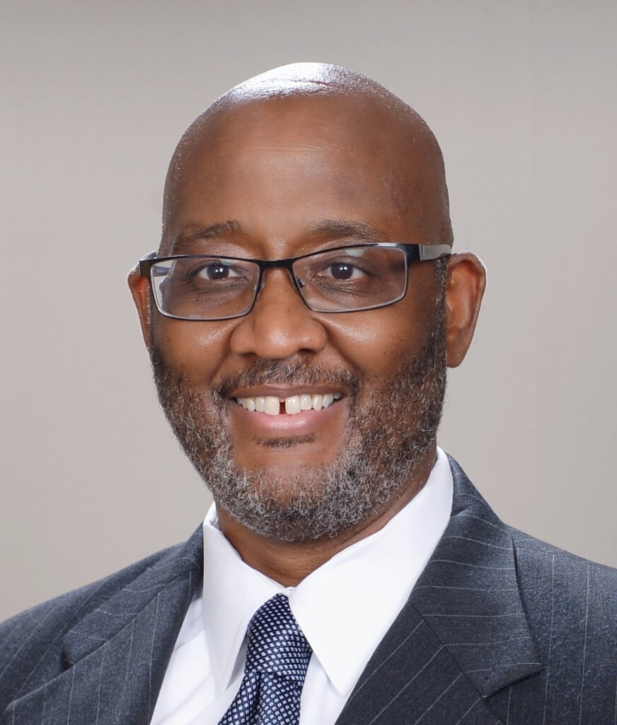 A man wearing glasses, a suit, and a tie, smiling at the camera against a plain, light-colored background.