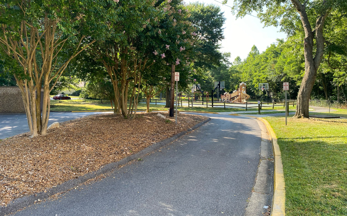 Paved path curves past trees and a mulched area toward a playground with slides and swings, bordered by grass and shaded by tall trees.