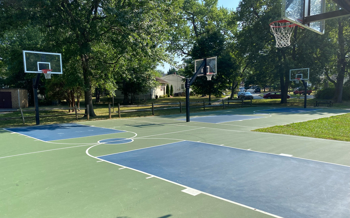 Outdoor basketball court with four hoops, surrounded by trees and houses on a sunny day. Shadows from trees fall across the court.