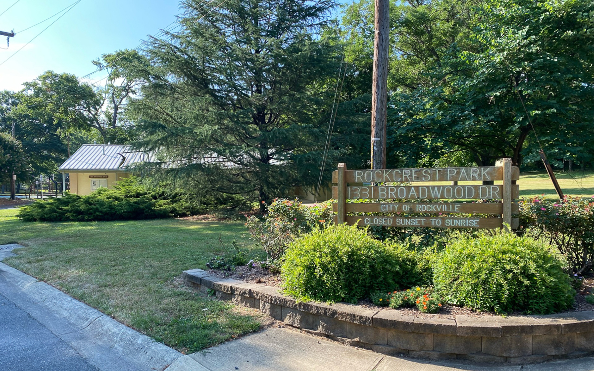 A wooden sign for Rockcrest Park at 1311 Broadwood Dr. stands near a lawn, trees, and a yellow building in a residential area.