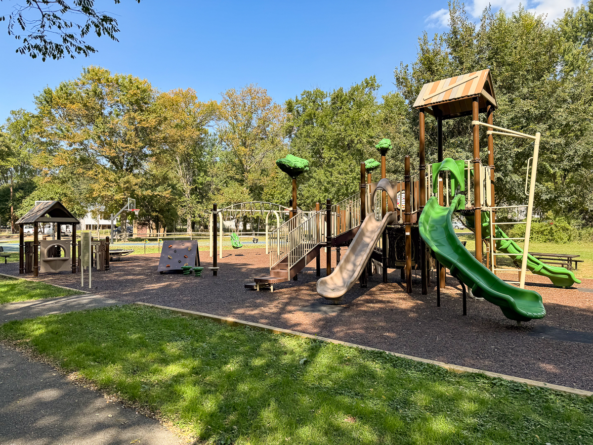 Outdoor playground with slides, climbing structures, and swings on a sunny day, surrounded by trees and grass. No people present.