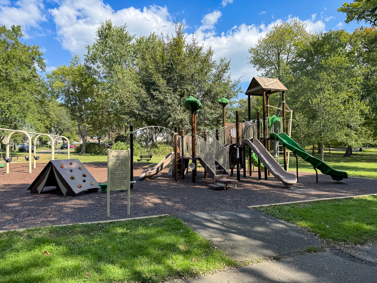A playground with slides, climbing structures, and swings is set in a park with green grass and trees under a partly cloudy sky.