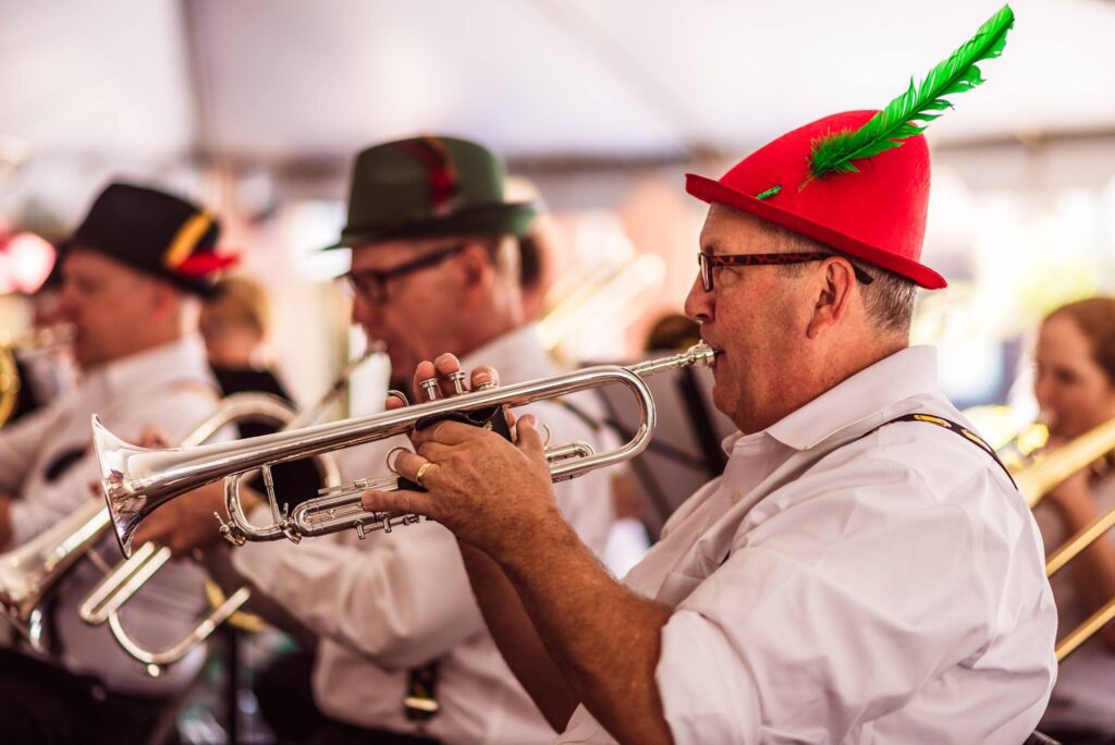 A group of musicians wearing traditional hats with feathers plays brass instruments at an event under a tent.