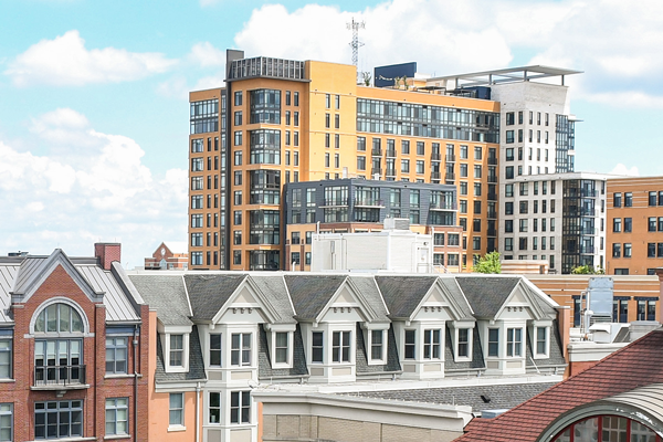 A modern multi-story apartment building stands behind older, gabled residential buildings under a partly cloudy sky.