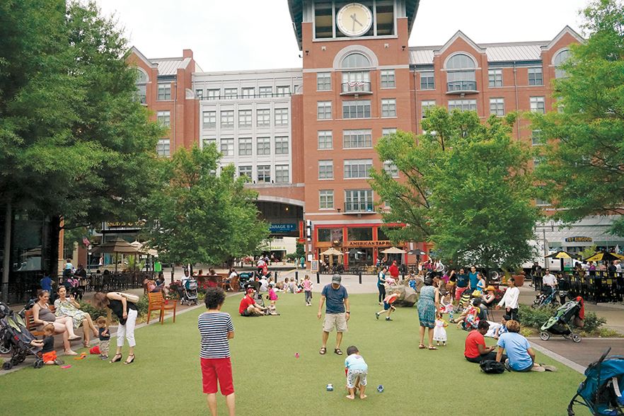 People, including children and adults, gather and play on a grassy area in an urban plaza with trees and a large brick building in the background.