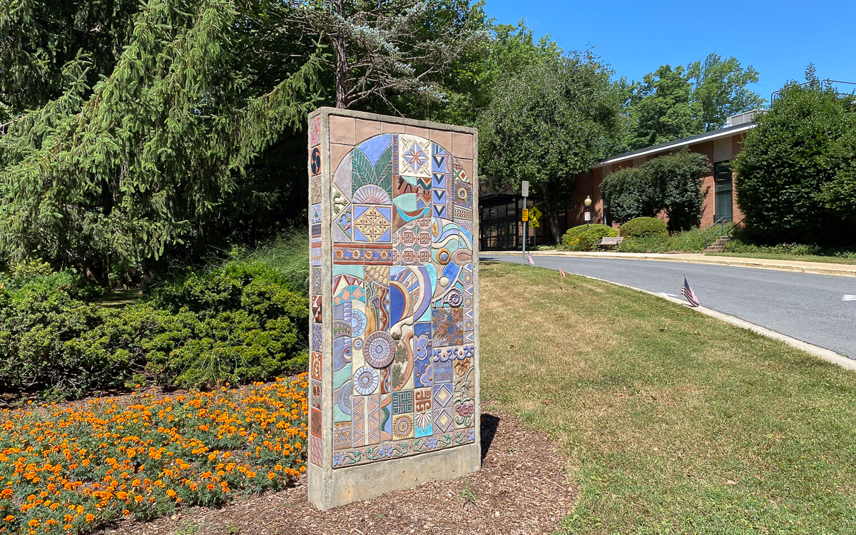 A colorful tile mural stands upright in a landscaped area with flowers and grass near a driveway and a brick building in the background.