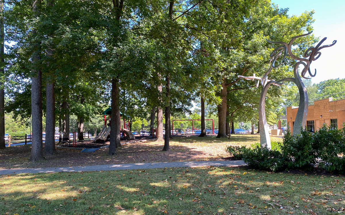 A shaded park area with tall trees, a playground in the background, a modern metal sculpture to the right, and a brick building on the far right.