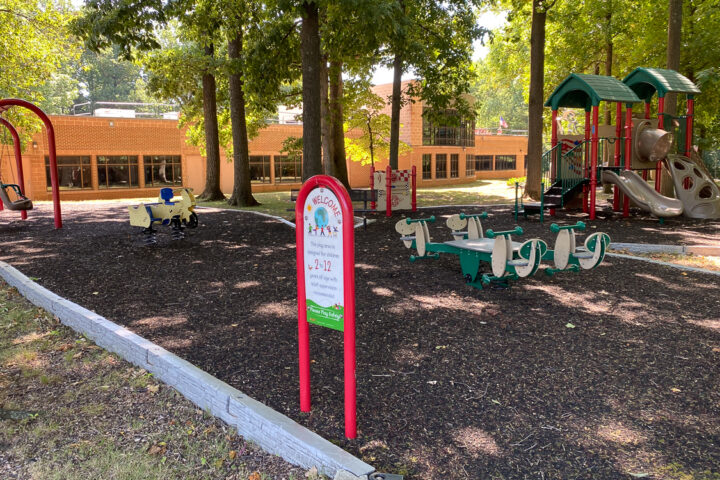 A playground with animal-shaped spring riders, a sign with park rules, and a play structure with slides, surrounded by trees and mulch, next to a brick building.