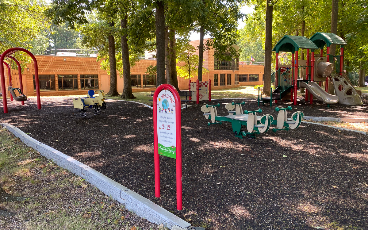 A playground with animal-shaped spring riders, a sign with park rules, and a play structure with slides, surrounded by trees and mulch, next to a brick building.