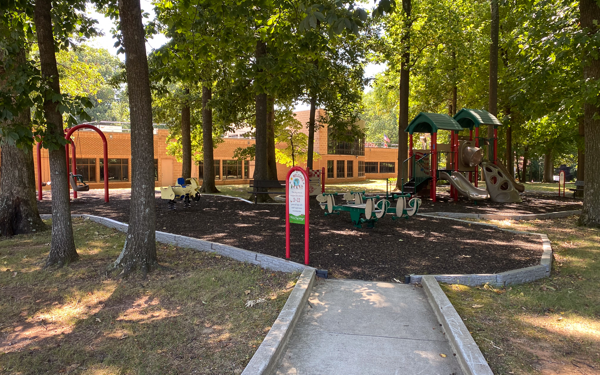 A small playground with swings, a seesaw, and climbing equipment surrounded by trees, located in front of a brick building.