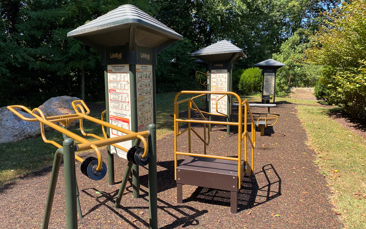 Outdoor fitness equipment and instructional signs on a mulch path in a park, surrounded by trees and greenery on a sunny day.