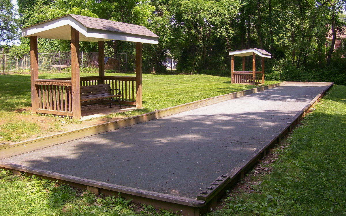 A bocce ball court in a grassy park area with two wooden benches under small shelters along the side, surrounded by trees.