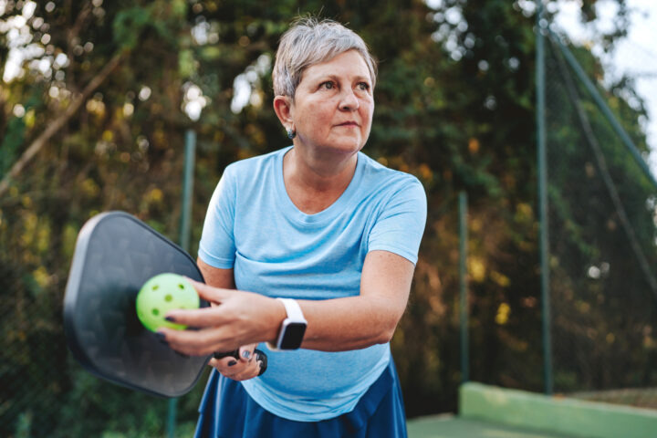 Older woman in athletic wear prepares to serve a pickleball on an outdoor court, holding a paddle and a ball, with trees and a fence in the background.