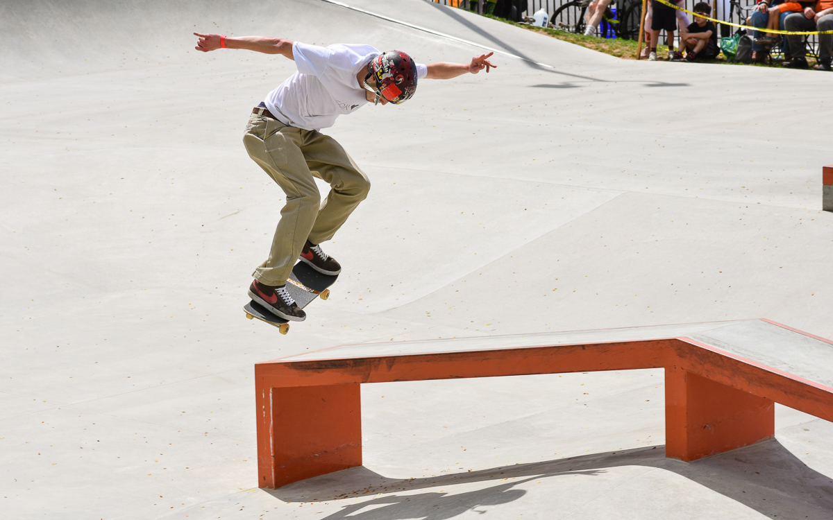 A skateboarder wearing a helmet and white shirt performs a trick over a red rail at an outdoor skate park.