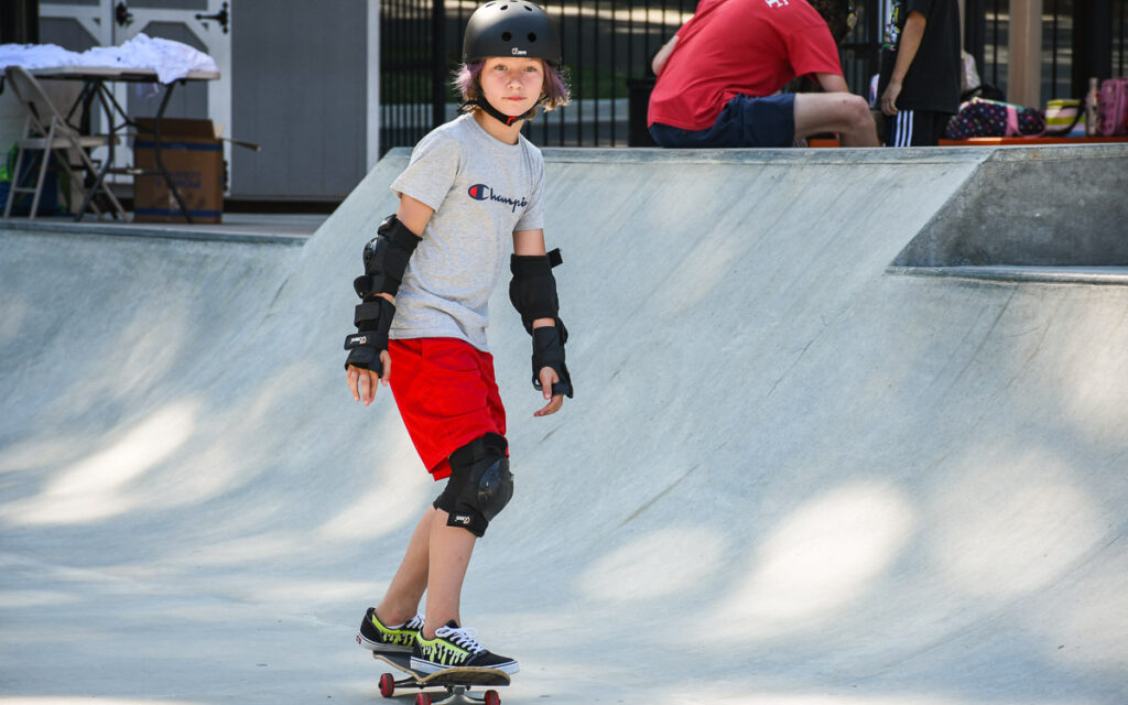 Young person wearing a helmet and protective gear skateboards in a concrete skate park during the day.