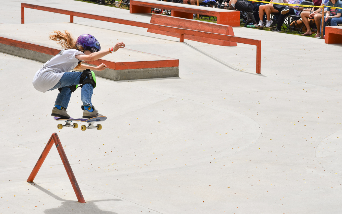 A skateboarder wearing a helmet performs a jump over a metal rail at an outdoor skatepark, with spectators watching in the background.