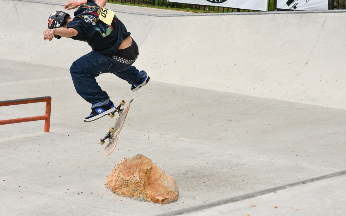 A skateboarder performs a trick over a rock in a concrete skatepark, with the skateboard mid-air and the skater wearing a helmet and loose clothing.