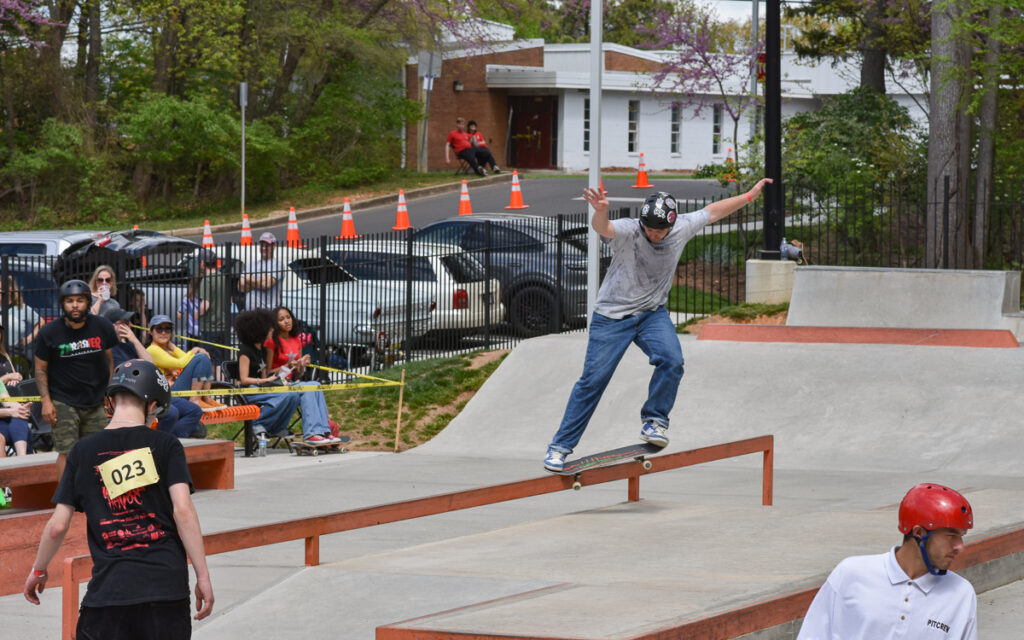 A skateboarder in a gray shirt and helmet grinds on a rail at an outdoor skate park while others watch and participate nearby.